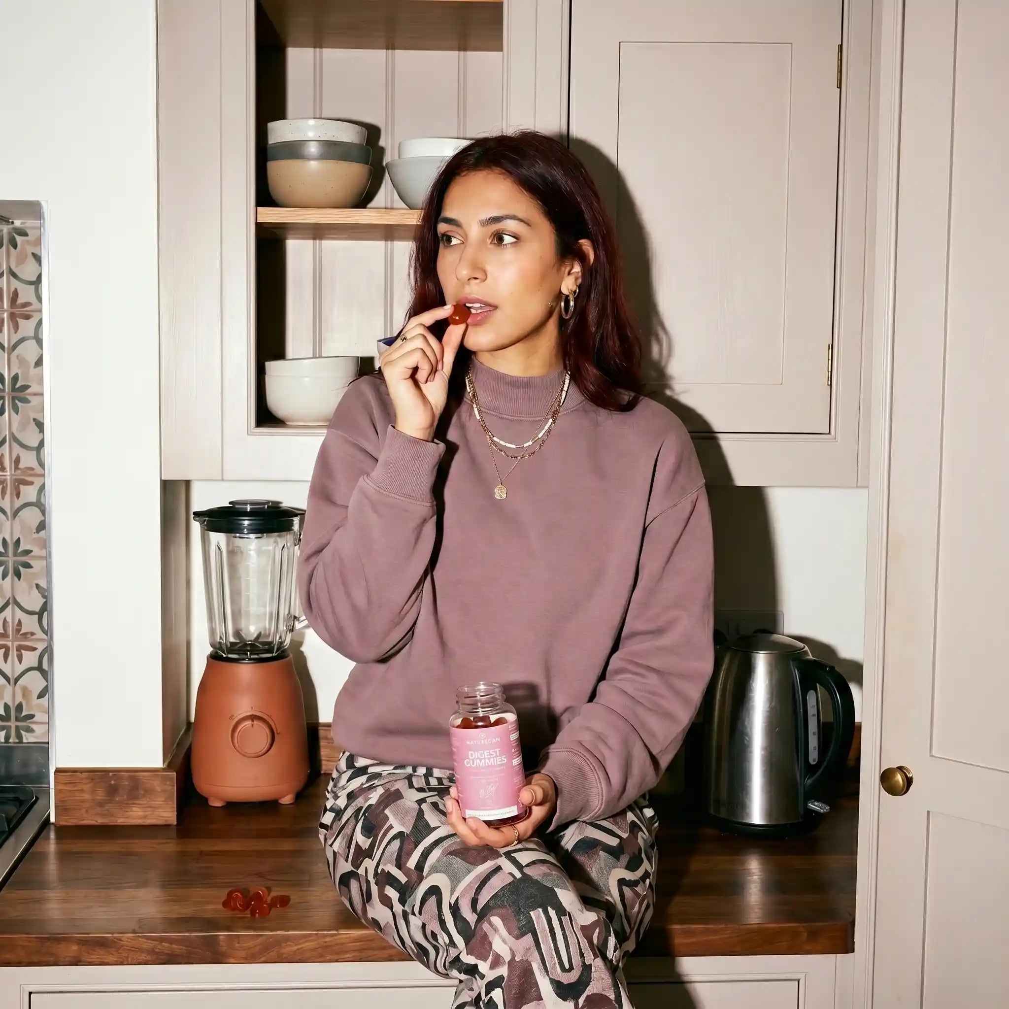Woman sitting at a kitchen counter with a pink jar, surrounded by kitchen appliances and decor.