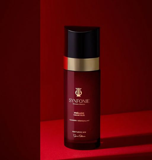 A bottle of Synfonie Prélude by Naturecan from Synergie Skincare, featuring a deep burgundy body, a shiny gold band, and a black cap, set against a matching dark red and black background.